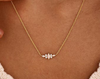 May include: A gold chain necklace featuring a central pendant composed of five small, marquise-cut white stones arranged in a cluster. The delicate metal chain connects to each side of the stone setting, creating a simple, balanced jewelry piece.