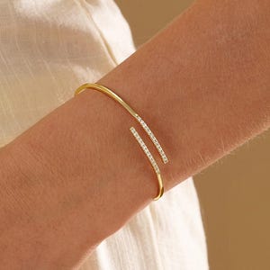 May include: A gold-toned open cuff bracelet featuring two parallel, slightly overlapping ends. Each end is accented with a row of small, clear, sparkling stones set along the outer surface of the thin, polished metal band.