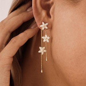 May include: Gold-coloured drop earrings with three delicate, white flower pendants. The earrings have a linear design with small, dangling accents. The floral design adds a touch of elegance to the jewellery.