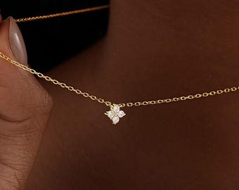 May include: A delicate gold necklace featuring a small, four-petal flower pendant with sparkling white gemstones. The necklace is held up, showcasing its fine chain and the intricate detail of the floral design. The image is a close-up, highlighting the jewelry's elegance.