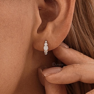 May include: Small gold hoop earrings with three clear, sparkling stones. The earrings are worn on the ear of a person with blonde hair. The person's hand is touching the earring.