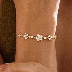 May include: A gold-toned bracelet featuring a floral and leaf design with clear gemstones. The bracelet is worn on a wrist, showcasing its delicate details and elegant style. The bracelet's design includes a central flower and leaf accents.