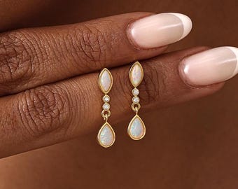 May include: A pair of gold drop earrings featuring a marquise-shaped opal at the top, two small round clear stones in the center, and a pear-shaped opal at the bottom. The earrings are suspended from a thin gold chain link design.