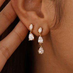 May include: Gold-toned earrings with three pear-shaped, clear gemstones dangling from each earring. The earrings are being worn, with one earring in focus and the other slightly out of focus. The background is a neutral color.
