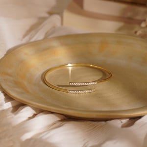 Puede incluir: Un brazalete abierto de tono dorado con dos extremos paralelos, cada uno adornado con una hilera de peque&ntilde;as piedras claras y brillantes. El brazalete tiene un dise&ntilde;o de banda delgada y elegante que se curva en forma circular con un acabado met&aacute;lico pulido.