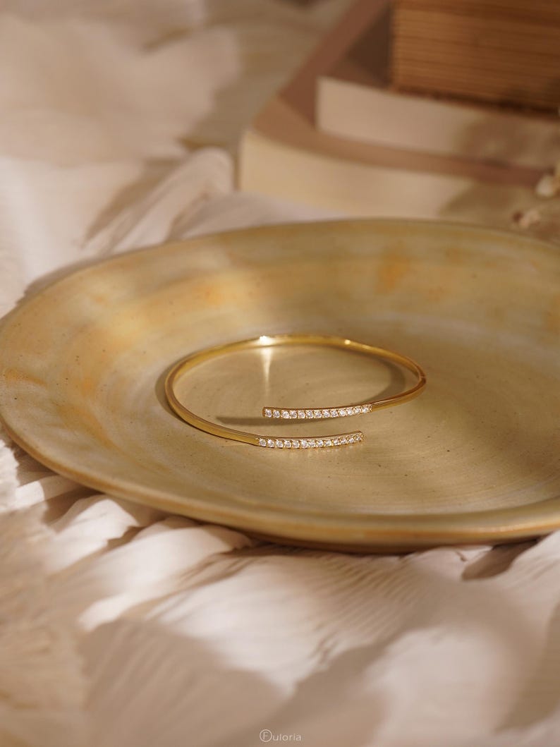 Puede incluir: Un brazalete abierto de tono dorado con dos extremos paralelos, cada uno adornado con una hilera de peque&ntilde;as piedras claras y brillantes. El brazalete tiene un dise&ntilde;o de banda delgada y elegante que se curva en forma circular con un acabado met&aacute;lico pulido.