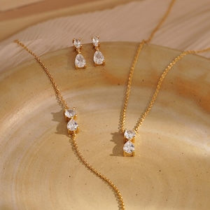 May include: A gold-toned jewelry set featuring a necklace, bracelet, and earrings. Each piece showcases clear, pear-shaped gemstones. The jewelry is displayed on a neutral-toned surface, with soft lighting enhancing the sparkle of the stones.