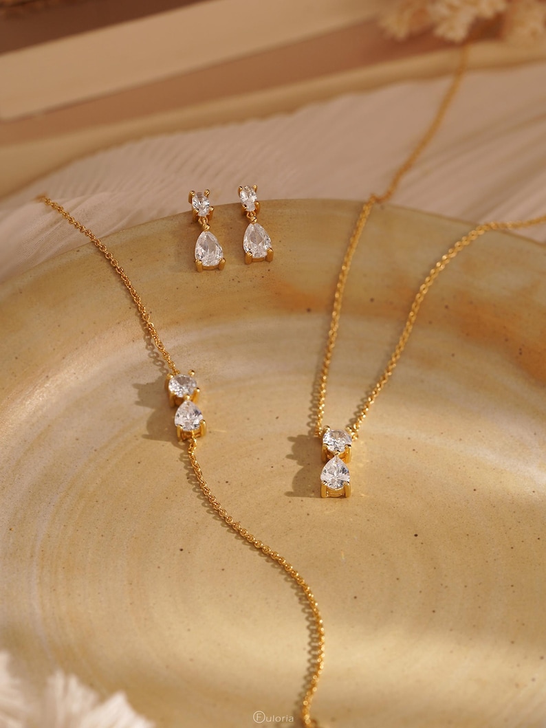 May include: A gold-toned jewelry set featuring a necklace, bracelet, and earrings. Each piece showcases clear, pear-shaped gemstones. The jewelry is displayed on a neutral-toned surface, with soft lighting enhancing the sparkle of the stones.