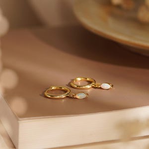 May include: A pair of gold hoop earrings, each featuring a small, marquise-shaped opal gemstone charm dangling from the bottom. The earrings have a polished metallic finish and a simple hinged closure mechanism, showcasing a delicate and refined design.