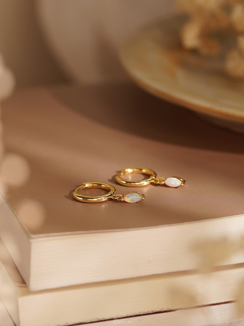 May include: A pair of gold hoop earrings, each featuring a small, marquise-shaped opal gemstone charm dangling from the bottom. The earrings have a polished metallic finish and a simple hinged closure mechanism, showcasing a delicate and refined design.