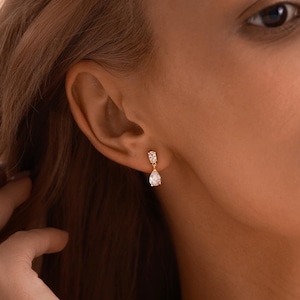 May include: Gold-toned drop earrings with a pear-shaped, clear gemstone. The earrings are set with smaller, clear stones at the top. The earrings are shown on a person's ear, with the focus on the jewelry. The brand name "Ouloria" is visible at the bottom.