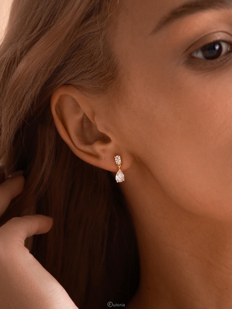 May include: Gold-toned drop earrings with a pear-shaped, clear gemstone. The earrings are set with smaller, clear stones at the top. The earrings are shown on a person's ear, with the focus on the jewelry. The brand name "Ouloria" is visible at the bottom.
