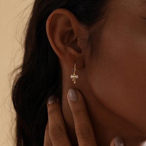 Puede incluir: Un pendiente de aro dorado con un peque&ntilde;o dije decorativo. El dije presenta un dise&ntilde;o en forma de lazo con peque&ntilde;os detalles brillantes. El pendiente se lleva en la oreja de una persona con cabello oscuro.