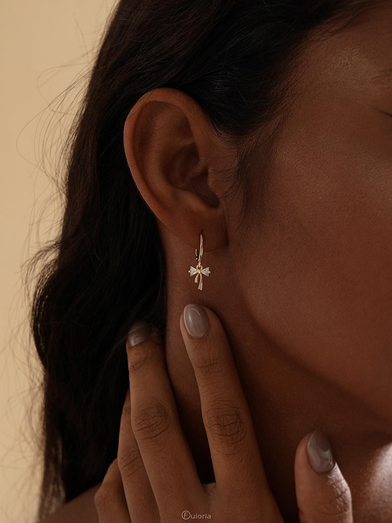 Puede incluir: Un pendiente de aro dorado con un peque&ntilde;o dije decorativo. El dije presenta un dise&ntilde;o en forma de lazo con peque&ntilde;os detalles brillantes. El pendiente se lleva en la oreja de una persona con cabello oscuro.