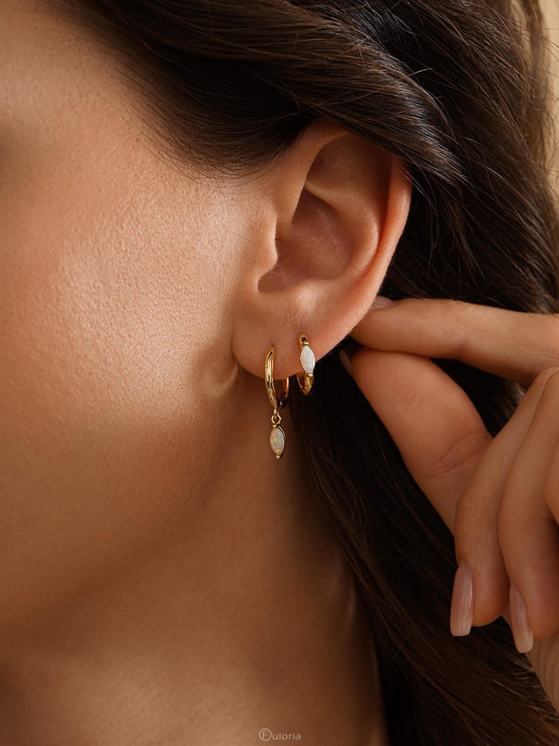 May include: Two gold hoop earrings worn in the earlobe. The smaller hoop features a marquise-cut white opal stone set directly into the band. The larger hoop hangs below it and features a matching marquise-cut white opal charm dangling from the base.