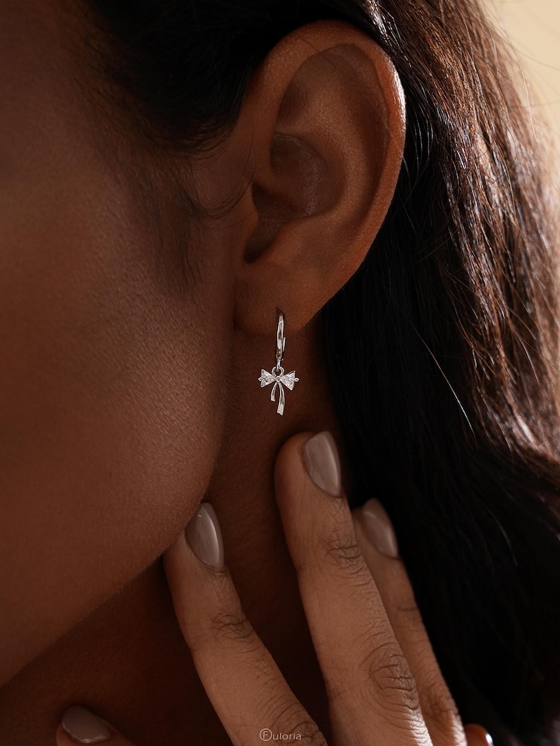 Puede incluir: Un pendiente plateado en forma de lazo con un aro peque&ntilde;o. El pendiente cuelga cerca de la oreja de una persona. El lazo tiene un peque&ntilde;o acento brillante en el centro. Una joya delicada.