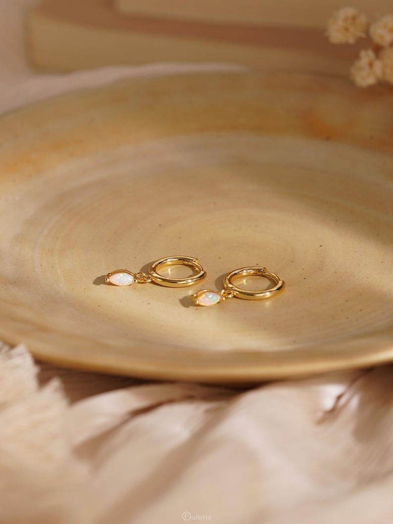 May include: A pair of gold hoop earrings, each featuring a small, dangling marquise-cut opal gemstone. The earrings have a polished metallic finish and a secure hinged closure mechanism, showcasing a simple and refined design for everyday wear.