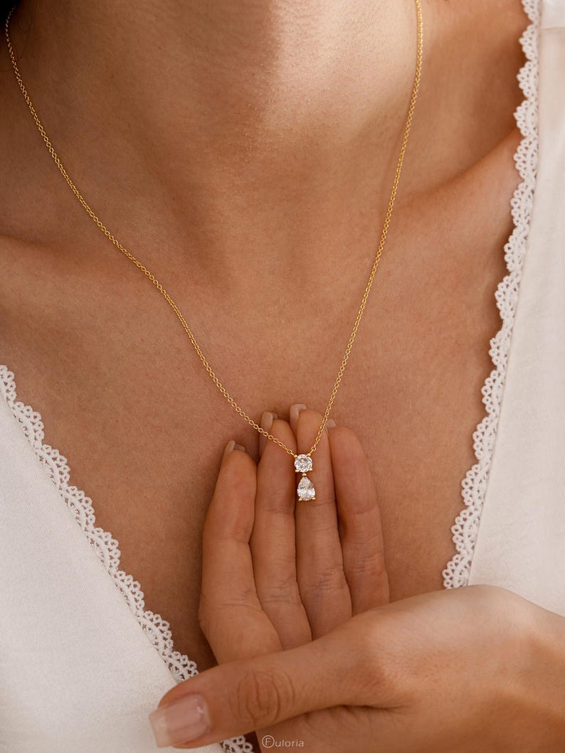 May include: A delicate gold necklace featuring two diamond pendants: a square-cut stone above a pear-shaped stone. The necklace is displayed against a white background. The jewelry is from the brand "Fuloria".