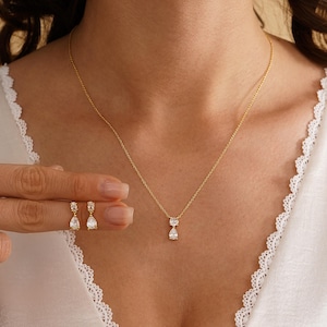 May include: A gold-colored necklace and matching earrings set. The necklace features a delicate chain with a pear-shaped pendant. The earrings are also pear-shaped and dangle from the ear. The jewelry is displayed on a person wearing a white top.