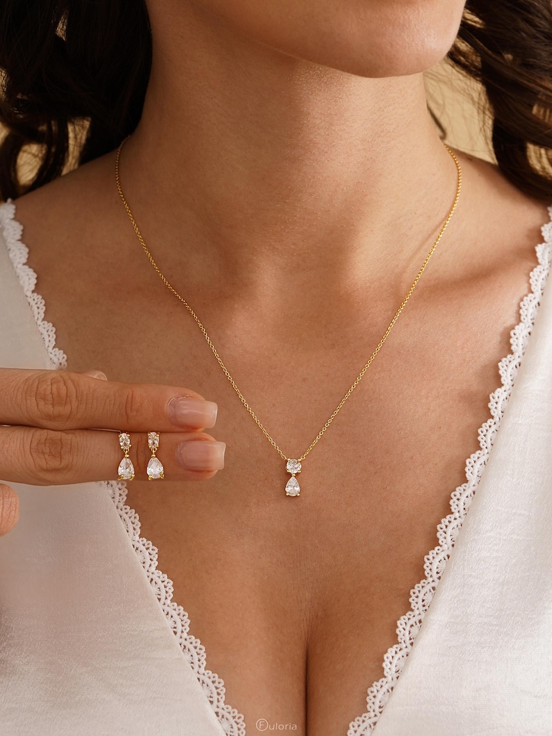 May include: A gold-colored necklace and matching earrings set. The necklace features a delicate chain with a pear-shaped pendant. The earrings are also pear-shaped and dangle from the ear. The jewelry is displayed on a person wearing a white top.