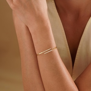 Puede incluir: Una pulsera abierta de tono dorado con dos barras paralelas que se superponen ligeramente. Cada barra est&aacute; decorada con una fila de peque&ntilde;as piedras claras y brillantes en la parte superior. La pulsera tiene un dise&ntilde;o de metal pulido.