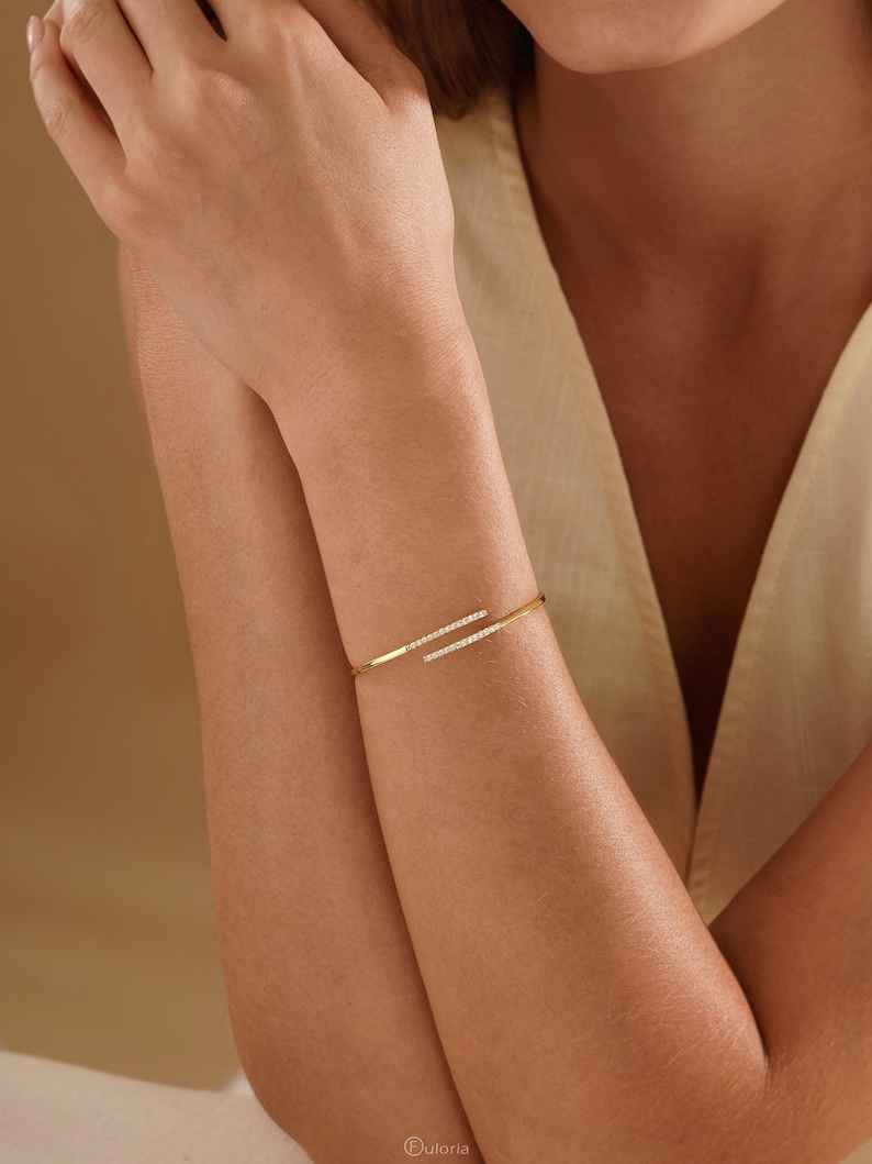 Puede incluir: Una pulsera abierta de tono dorado con dos barras paralelas que se superponen ligeramente. Cada barra est&aacute; decorada con una fila de peque&ntilde;as piedras claras y brillantes en la parte superior. La pulsera tiene un dise&ntilde;o de metal pulido.