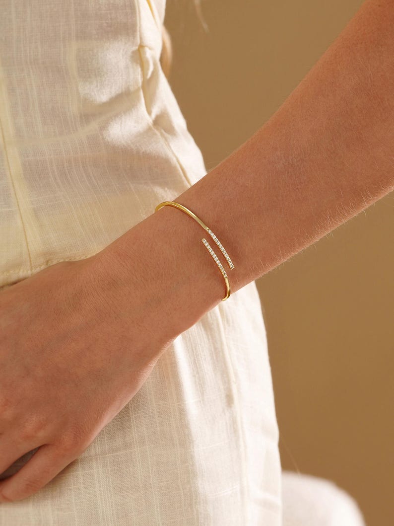 Puede incluir: Una pulsera abierta de tono dorado con dos extremos paralelos que se superponen ligeramente. Cada extremo est&aacute; decorado con una hilera de peque&ntilde;as piedras claras y brillantes incrustadas en la superficie exterior de la fina banda de metal pulido.
