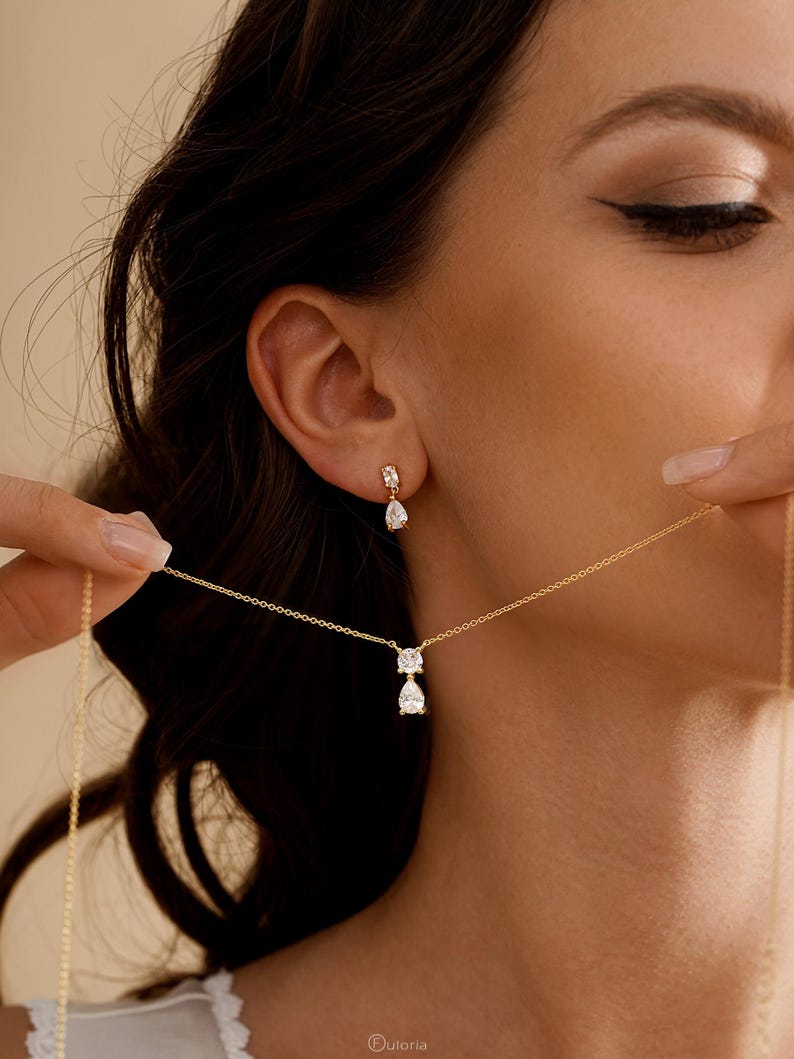 May include: A gold necklace and matching earrings set. The necklace features a pendant with two clear, teardrop-shaped stones. The earrings have a similar design, with a single teardrop stone. The jewelry is displayed against a soft, neutral background.