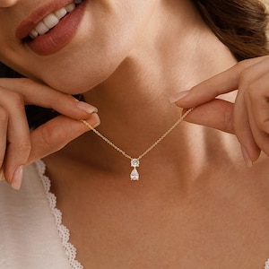 May include: A delicate gold necklace featuring two clear, sparkling gemstones. The top stone is round, and the bottom stone is a teardrop shape. The necklace is displayed against a soft, neutral background, highlighting the jewelry's elegance.