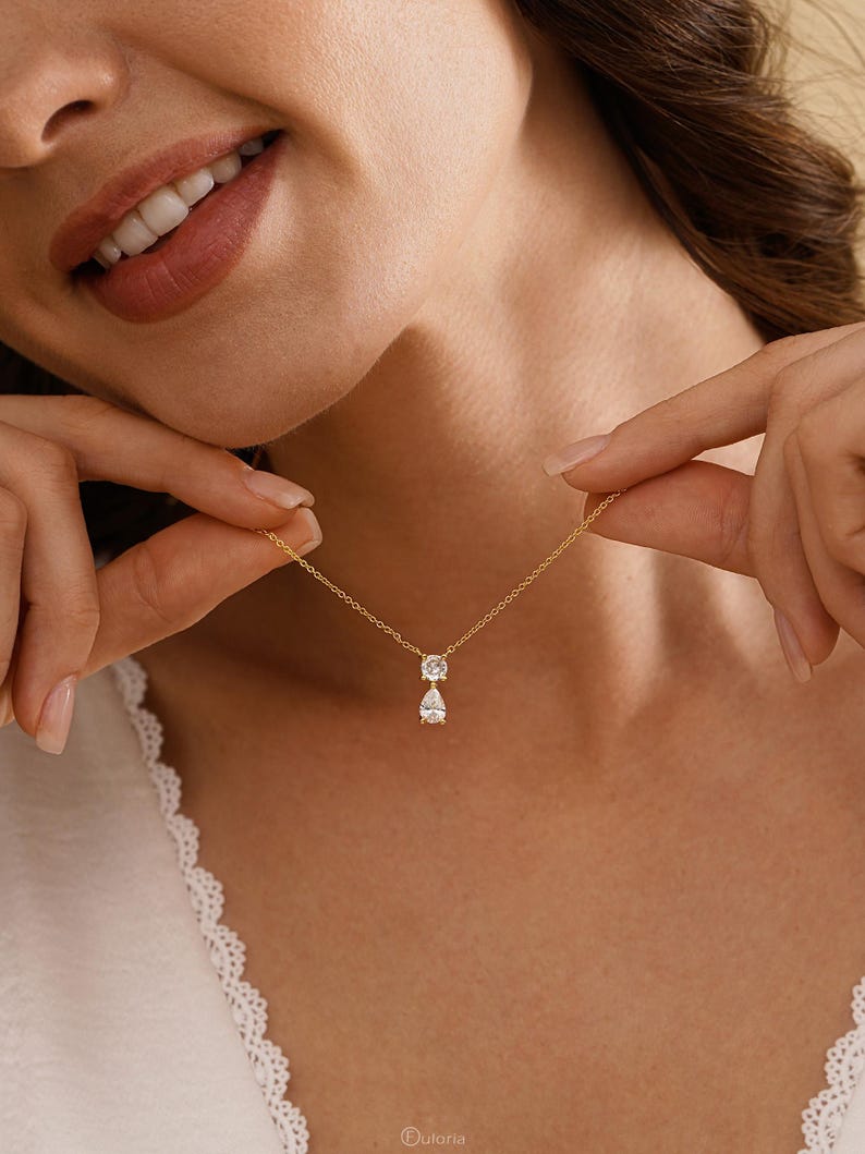 May include: A delicate gold necklace featuring two clear, sparkling gemstones. The top stone is round, and the bottom stone is a teardrop shape. The necklace is displayed against a soft, neutral background, highlighting the jewelry's elegance.