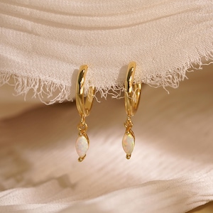 May include: A pair of gold hoop earrings featuring dangling marquise-cut opal charms. Each earring consists of a polished gold hoop with a small, iridescent white opal stone suspended from the bottom. The jewelry displays a bright, reflective finish.