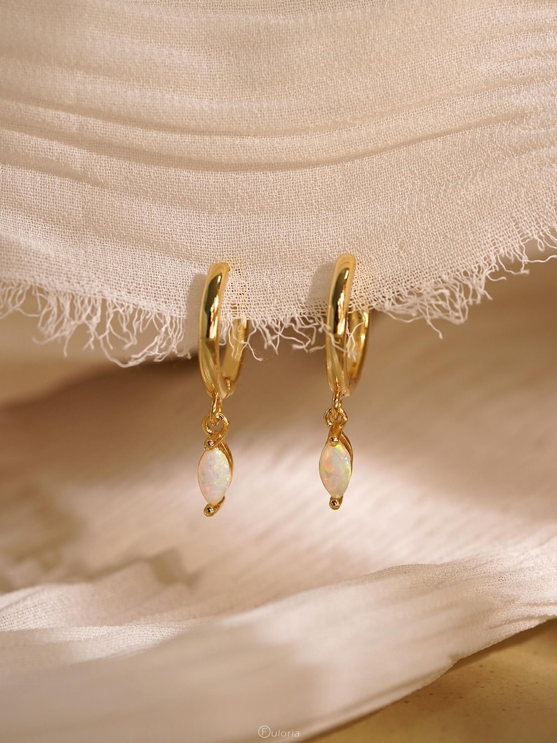 May include: A pair of gold hoop earrings featuring dangling marquise-cut opal charms. Each earring consists of a polished gold hoop with a small, iridescent white opal stone suspended from the bottom. The jewelry displays a bright, reflective finish.