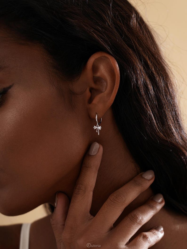 Puede incluir: Primer plano de una oreja con un peque&ntilde;o pendiente de aro plateado con un dije en forma de lazo. El pendiente est&aacute; detallado con peque&ntilde;os acentos brillantes. La persona tiene un tono de piel medio y cabello oscuro. El fondo es de un color suave y neutro.