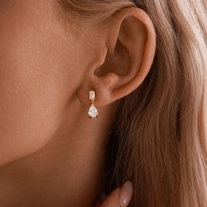 May include: A close-up of a gold earring with a rectangular top and a teardrop-shaped gem. The earring is worn on an ear, with the focus on the jewelry's design and the ear's shape. The earring is a delicate piece of jewelry.