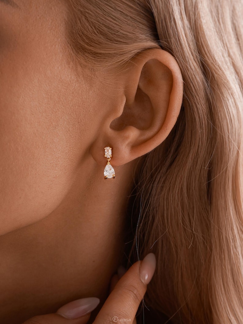 May include: A close-up of a gold earring with a rectangular top and a teardrop-shaped gem. The earring is worn on an ear, with the focus on the jewelry's design and the ear's shape. The earring is a delicate piece of jewelry.
