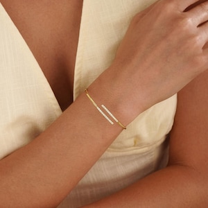Puede incluir: Una pulsera de brazalete abierto en tono dorado con dos barras paralelas, cada una decorada con una hilera de peque&ntilde;as piedras claras y brillantes. La pulsera tiene un dise&ntilde;o elegante con un acabado pulido, creando un aspecto refinado en la mu&ntilde;eca.