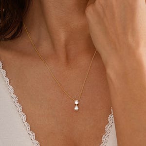 May include: A delicate gold necklace featuring two clear, sparkling gemstones: a round cut and a pear cut. The pendant hangs from a fine gold chain. The necklace is displayed on a person's chest.