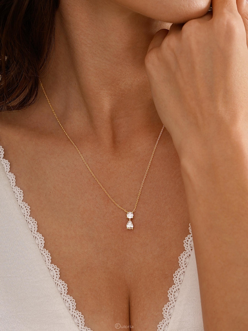 May include: A delicate gold necklace featuring two clear, sparkling gemstones: a round cut and a pear cut. The pendant hangs from a fine gold chain. The necklace is displayed on a person's chest.