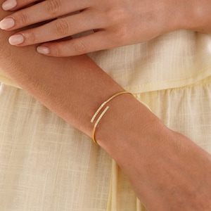 Puede incluir: Una pulsera abierta de tono dorado con dos barras paralelas ligeramente curvadas en los extremos, cada una incrustada con una hilera de peque&ntilde;as piedras claras y brillantes. La pulsera tiene una banda de metal pulido y fina.