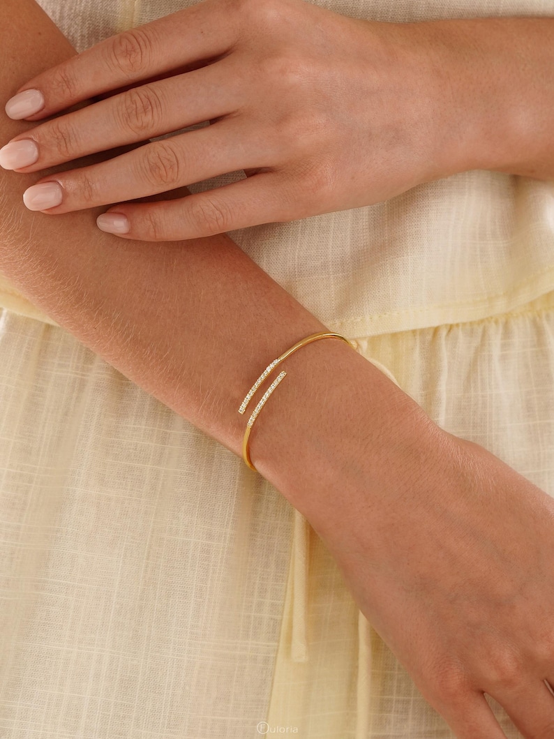 Puede incluir: Una pulsera abierta de tono dorado con dos barras paralelas ligeramente curvadas en los extremos, cada una incrustada con una hilera de peque&ntilde;as piedras claras y brillantes. La pulsera tiene una banda de metal pulido y fina.