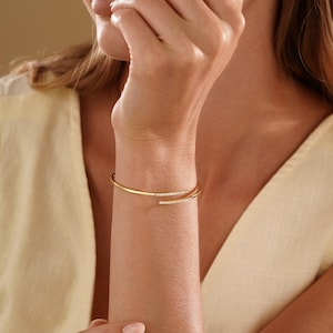 Puede incluir: Una pulsera tipo brazalete abierta en tono dorado con dos extremos, cada uno adornado con una hilera de peque&ntilde;as piedras claras y brillantes. La pulsera tiene un acabado liso y pulido con un dise&ntilde;o flexible que rodea la mu&ntilde;eca.