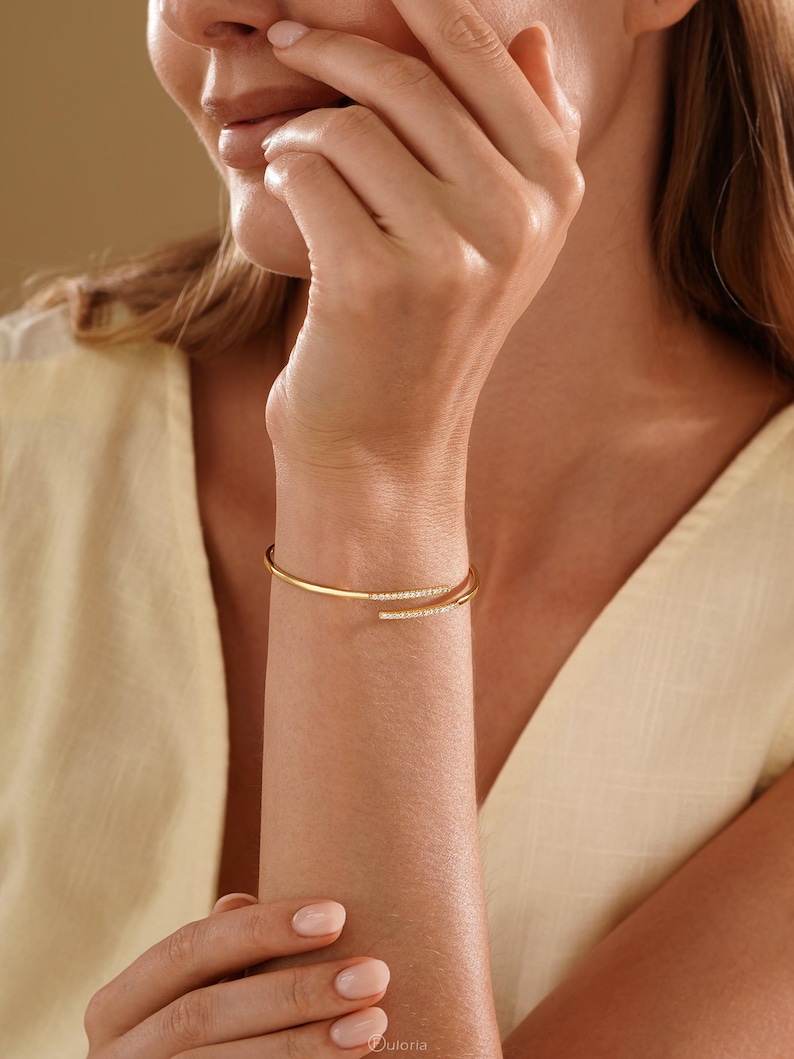 Puede incluir: Una pulsera tipo brazalete abierta en tono dorado con dos extremos, cada uno adornado con una hilera de peque&ntilde;as piedras claras y brillantes. La pulsera tiene un acabado liso y pulido con un dise&ntilde;o flexible que rodea la mu&ntilde;eca.