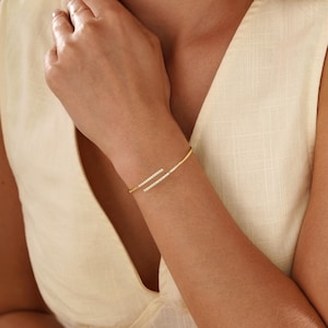 Puede incluir: Una pulsera de brazalete abierto en tono dorado con dos extremos paralelos ligeramente superpuestos. Cada extremo est&aacute; acentuado con una hilera de peque&ntilde;as piedras claras y brillantes. La pulsera tiene un dise&ntilde;o de banda de metal pulido.