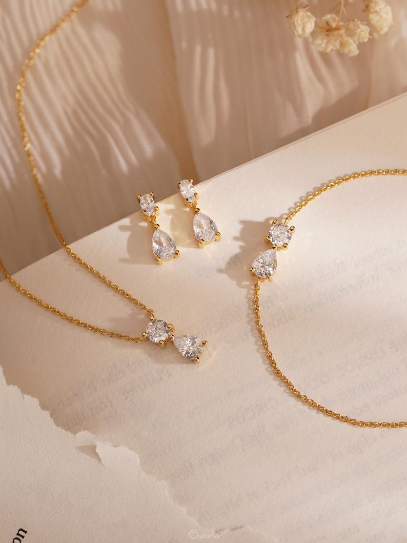 May include: A gold-toned jewelry set featuring a necklace, earrings, and bracelet. Each piece has a delicate chain and is adorned with clear, teardrop-shaped gemstones. The earrings dangle, and the necklace and bracelet have gemstones in a similar style.