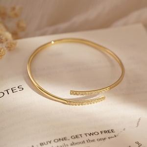 Puede incluir: Una pulsera abierta de tono dorado con dos extremos paralelos ligeramente superpuestos. Cada extremo est&aacute; adornado con una hilera de peque&ntilde;as piedras claras y brillantes. La pulsera tiene un acabado liso y pulido con un dise&ntilde;o circular flexible.