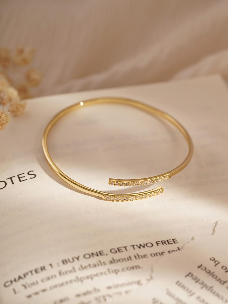 Puede incluir: Una pulsera abierta de tono dorado con dos extremos paralelos ligeramente superpuestos. Cada extremo est&aacute; adornado con una hilera de peque&ntilde;as piedras claras y brillantes. La pulsera tiene un acabado liso y pulido con un dise&ntilde;o circular flexible.