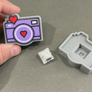 May include: A purple and grey camera-shaped mechanical keyboard keycap featuring a red heart on the lens and a red heart above the body, accompanied by a separate clear mechanical switch and a grey 3D-printed keycap base housing.