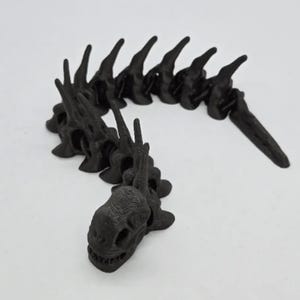 Puede incluir: Un juguete de dragón articulado negro, con un diseño esquelético detallado. La cabeza del dragón tiene cuencas oculares visibles y una sonrisa con dientes. El cuerpo está segmentado, lo que permite una postura flexible. El juguete está hecho de material negro mate.