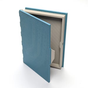 May include: A blue book-shaped box with a textured cover, partially open to reveal a canvas interior. The box has a light blue exterior and a neutral-colored interior, suggesting a storage or display item.