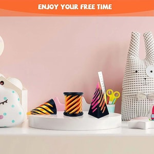 Könnte beinhalten: Eine dekorative Anordnung mit drei abstrakten Objekten mit schwarzen und orangen sowie rosa und orangen Designs. Die Objekte befinden sich auf einer weißen Plattform, mit einem rosa Hintergrund und dem Text "ENJOY YOUR FREE TIME" oben.